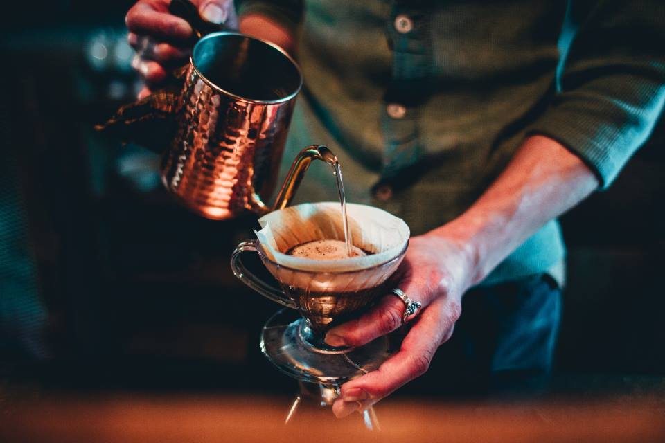 Un barista sirve agua caliente con una tetera de cobre sobre un filtro de vidrio