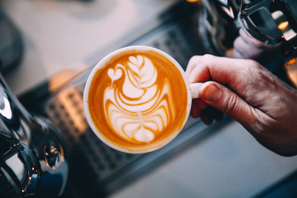 Una mano sostiene un latte con un cisne dibujado en la espuma, junto a la máquina de espresso